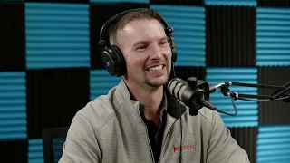 A person wearing headphones and a light-colored jacket smiles while speaking into a microphone in a soundproofed room.