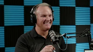 A man wearing headphones speaks into a microphone in a recording studio with blue and black soundproofing panels.