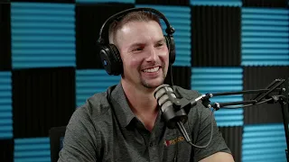 A man wearing headphones and a gray shirt speaks into a microphone while smiling, seated in front of a blue and black soundproofed wall.
.