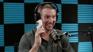 A man wearing headphones speaks into a microphone while holding a smartphone in his hand. He is in front of a background with blue and black acoustic panels.