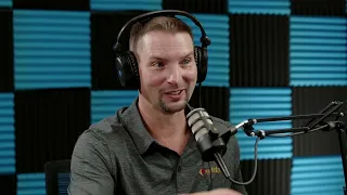 A man wearing headphones and speaking into a microphone in a soundproof room with blue and black acoustic panels.