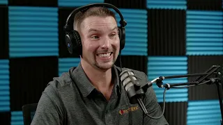 A man wearing headphones and a grey shirt with a logo smiles while speaking into a microphone in a soundproof room with black and blue foam panels.