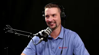 A man wearing headphones sits at a desk speaking into a microphone. He is dressed in a blue shirt and is in front of a black background.