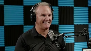 A person wearing headphones and speaking into a microphone, surrounded by a blue and black soundproofing wall.