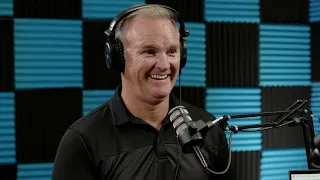 A person wearing headphones and smiling, speaking into a microphone in a sound-proofed studio.